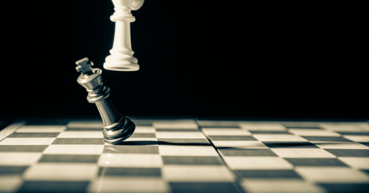 A black chess king toppling over beneath a white piece on a checkered board