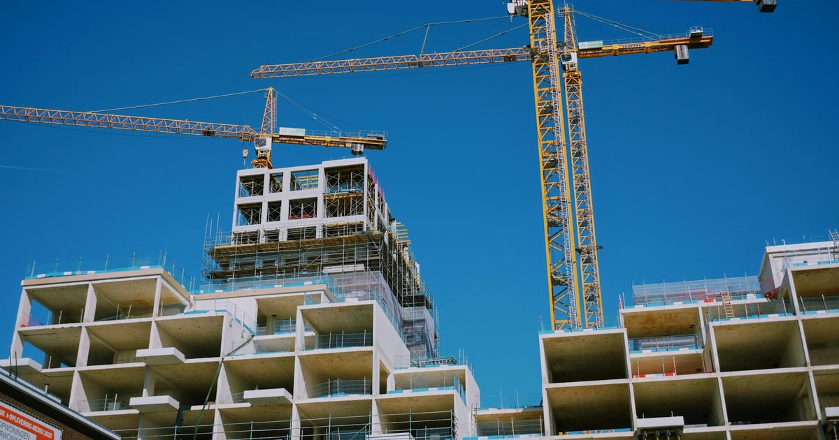 Tower cranes above a half-built apartment block with open concrete floors against a blue sky