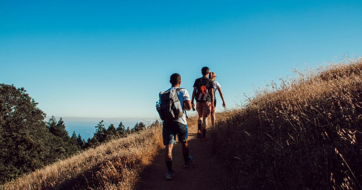 Two hikers with backpacks walking up a dry grassy ridge toward the sea